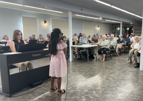 a young girl in a pink dress is singing at a Volunteer function hosted by the Vocal Academy to share their newly learned music and talent to the folks at the retirement home