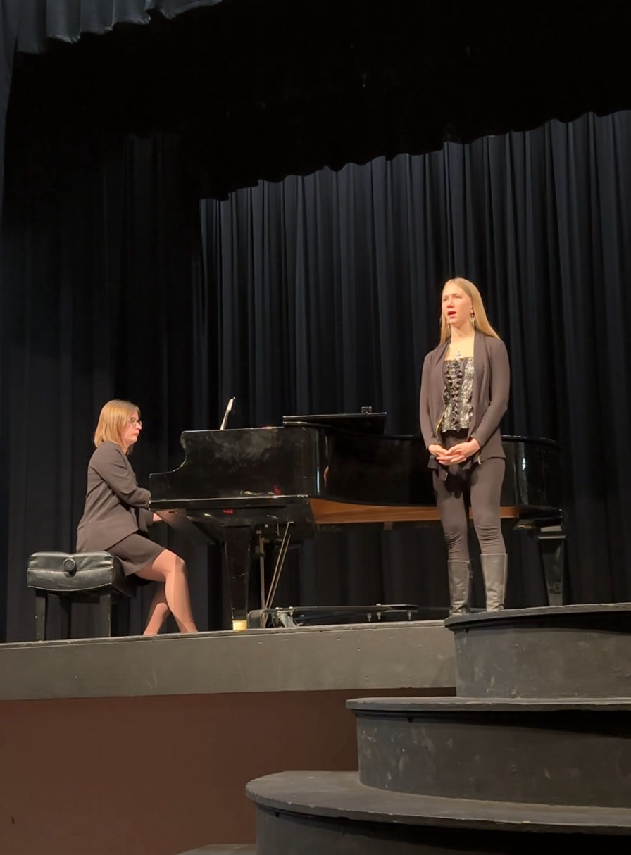 young student dressed in all black is performing on stage beside a grand piano demonstrating all she learned from her Singing Lessons Online
